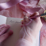 Bibliophile pen in the pastel pink and aqua Antoinette colour-way is held in a lace glove hand as it writes in the matching dot grid journal. The french inspired gel pen features floral motifs inspired by a classic literature aesthetic and 'BIBLIOPHILE' etched into the pen clip.