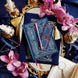 An overhead shot the blue literati bundle- a blue leather notebook with intricate gold foil detailing, accompanied by a matching blue pen, a bookmark, and a plush velvet book sleeve, all displayed on a surface with decorative elements like flowers and gold trim.