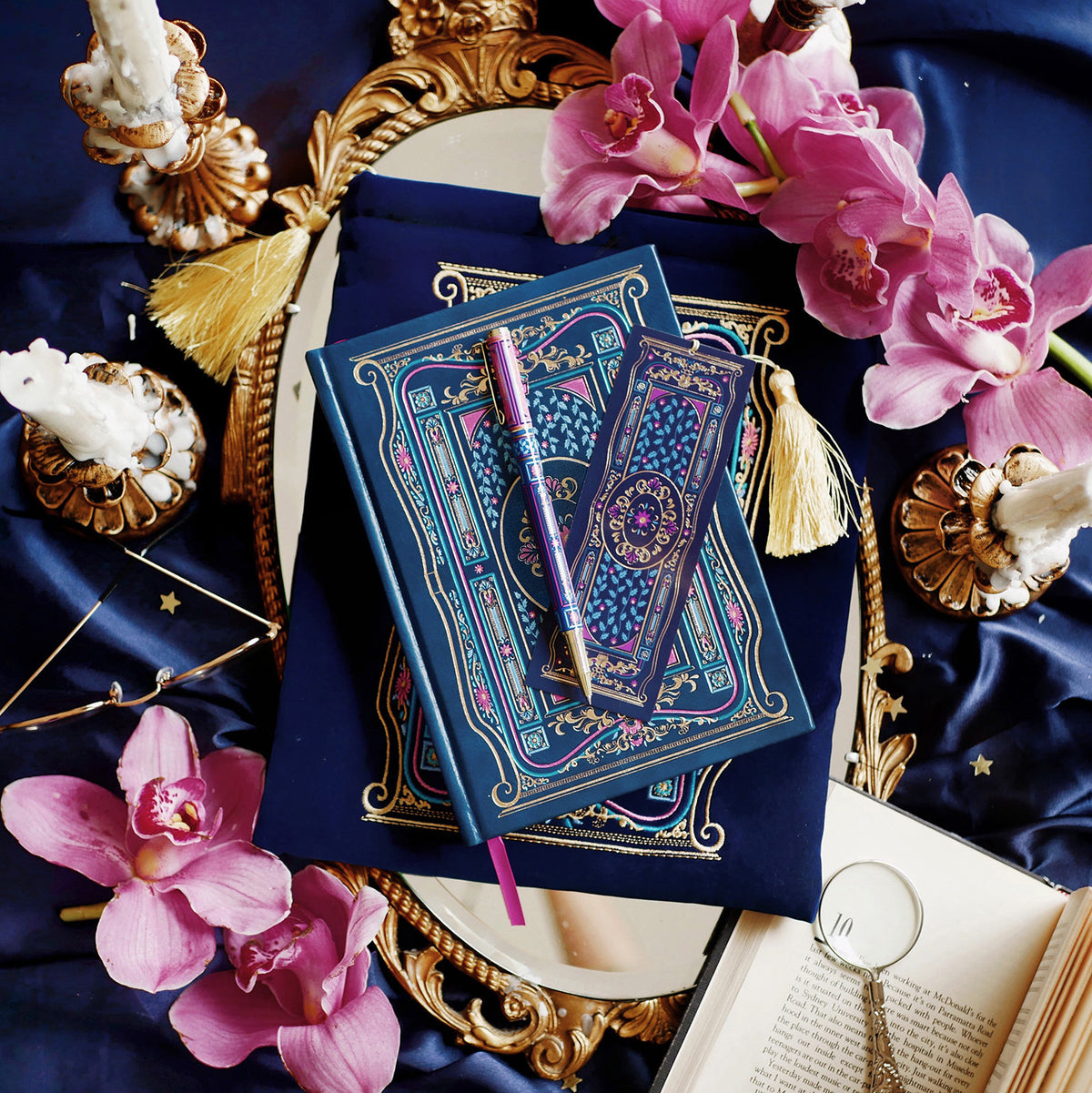 An overhead shot the blue literati bundle- a blue leather notebook with intricate gold foil detailing, accompanied by a matching blue pen, a bookmark, and a plush velvet book sleeve, all displayed on a surface with decorative elements like flowers and gold trim.