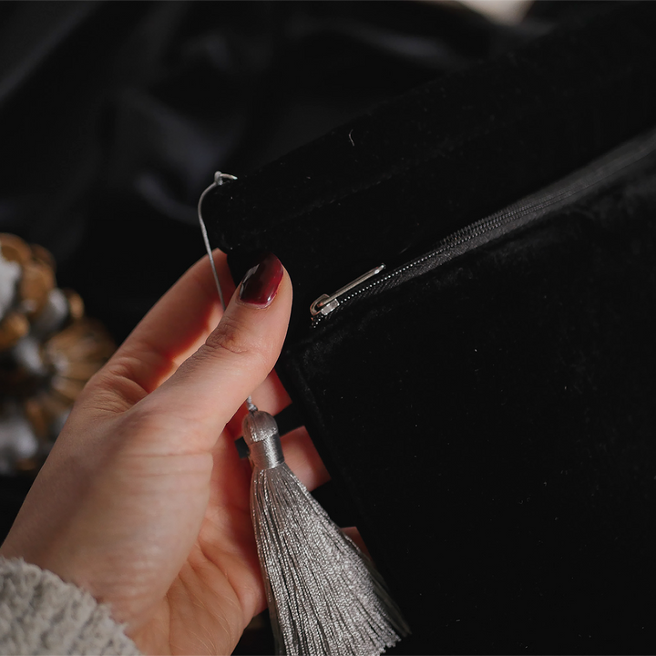 A detailed view of a hand holding the corner of a black velvet book sleeve, highlighting the silver zipper and tassel. The plush texture of the sleeve contrasts beautifully with the dark background