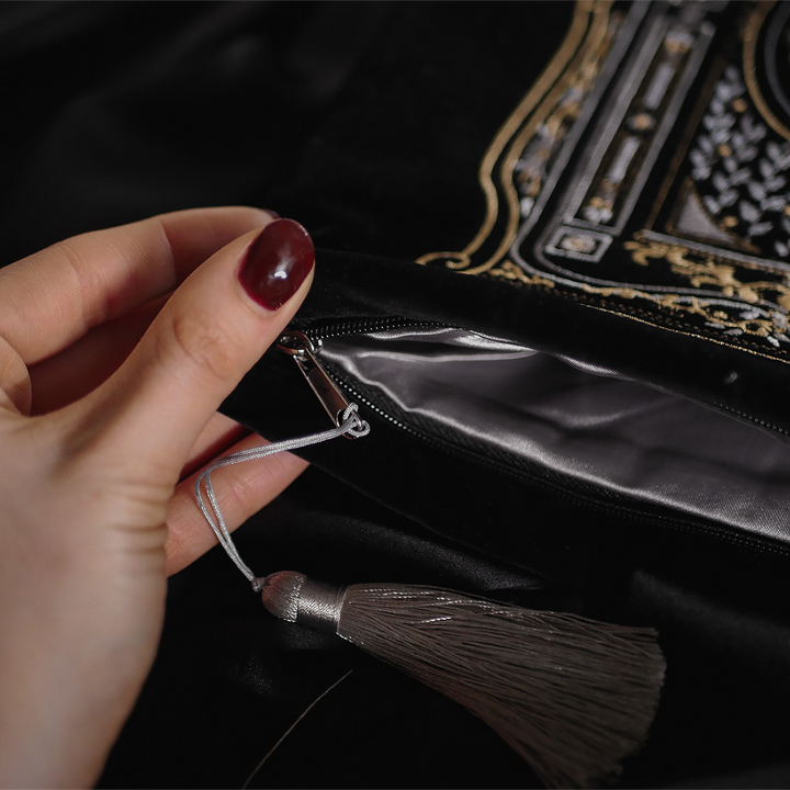 A detailed view of a hand holding the corner of a black velvet book sleeve, highlighting the silver zipper and tassel. The plush texture of the sleeve contrasts beautifully with the dark background