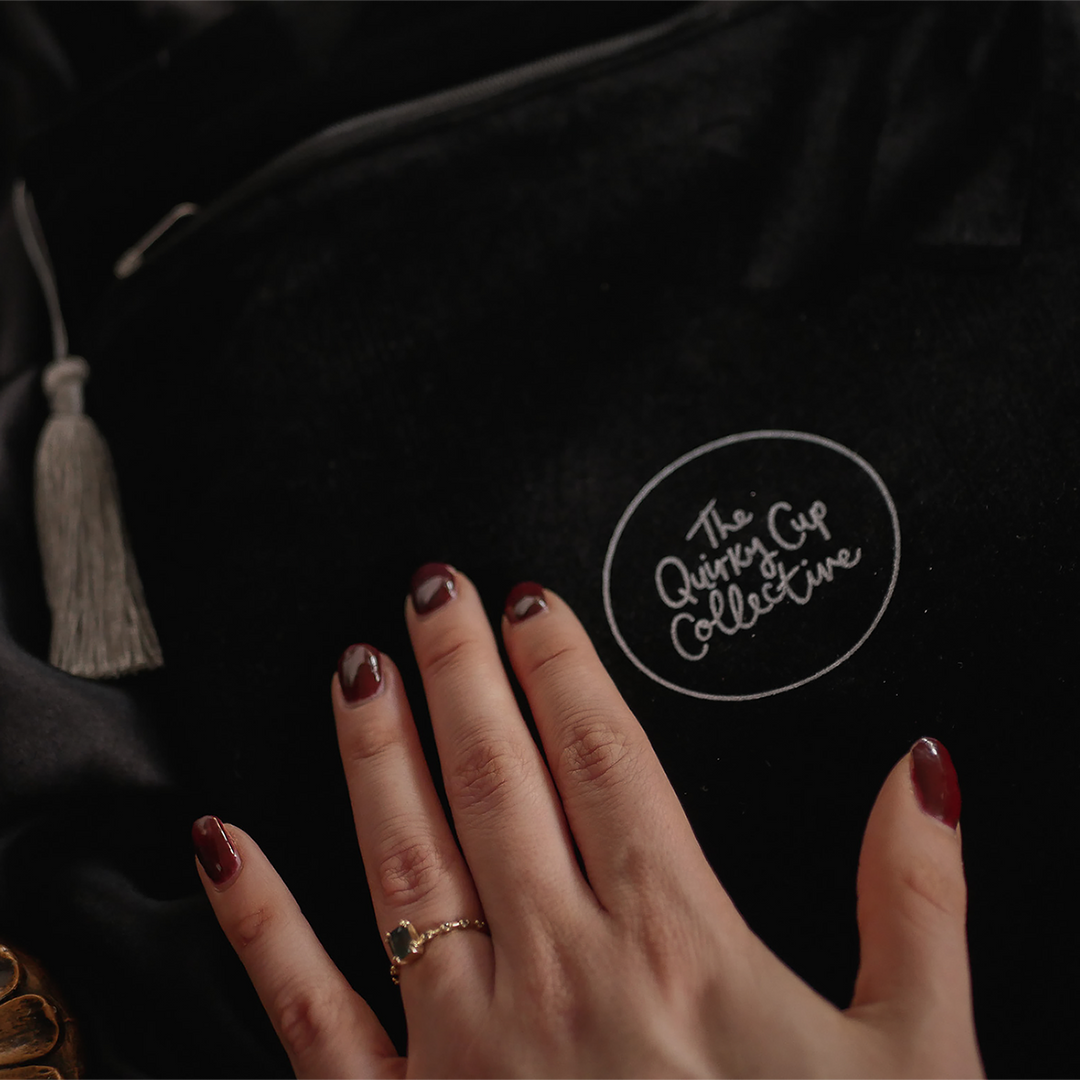  A close-up of a hand gently resting on a black velvet book sleeve embossed with "The Quirky Cup Collective" logo in white. The sleeve features a luxurious texture, paired with a silver tassel zipper pull.
