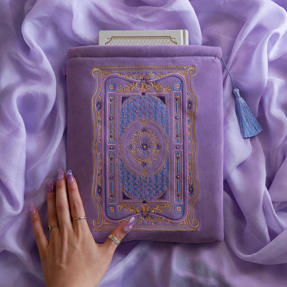 The soft lilac book sleeve as part of the classic book inspired Wisteria Literati collection sits on a light purple fabric background. This soft purple velvet book case features a slightly Victorian book inspired motif in shades of gold, white, pink and purple alongside intricate foil embroidery. The case features a soft light blue tassel.