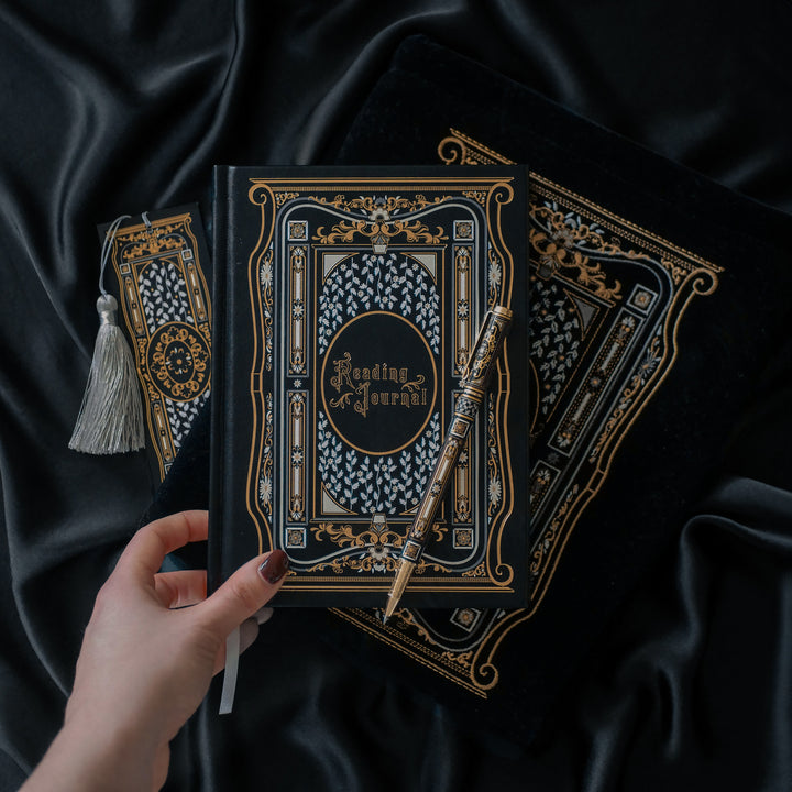 A hand holding a luxurious reading journal with an intricate gold and white design, accompanied by an ornate matching pen. The journal lies on top of a black velvet book sleeve from The Quirky Cup Collective, showcasing a coordinated design. A matching bookmark with a silver tassel is also partially visible. All items are set against a smooth black satin fabric background, creating an elegant and cohesive visual presentation.