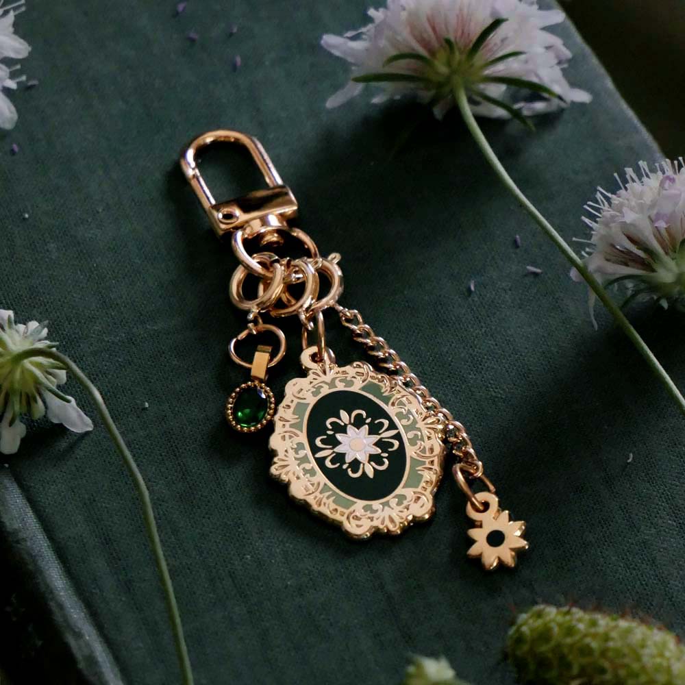 On a green fabric covered book surrounded by flowers sits the Sage green Literati charmlet. the gold clip accessory features detachable charms of a oval green flower motif, a green gem and a small gold flower.
