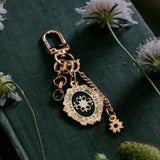 On a green fabric covered book surrounded by flowers sits the Sage green Literati charmlet. the gold clip accessory features detachable charms of a oval green flower motif, a green gem and a small gold flower.