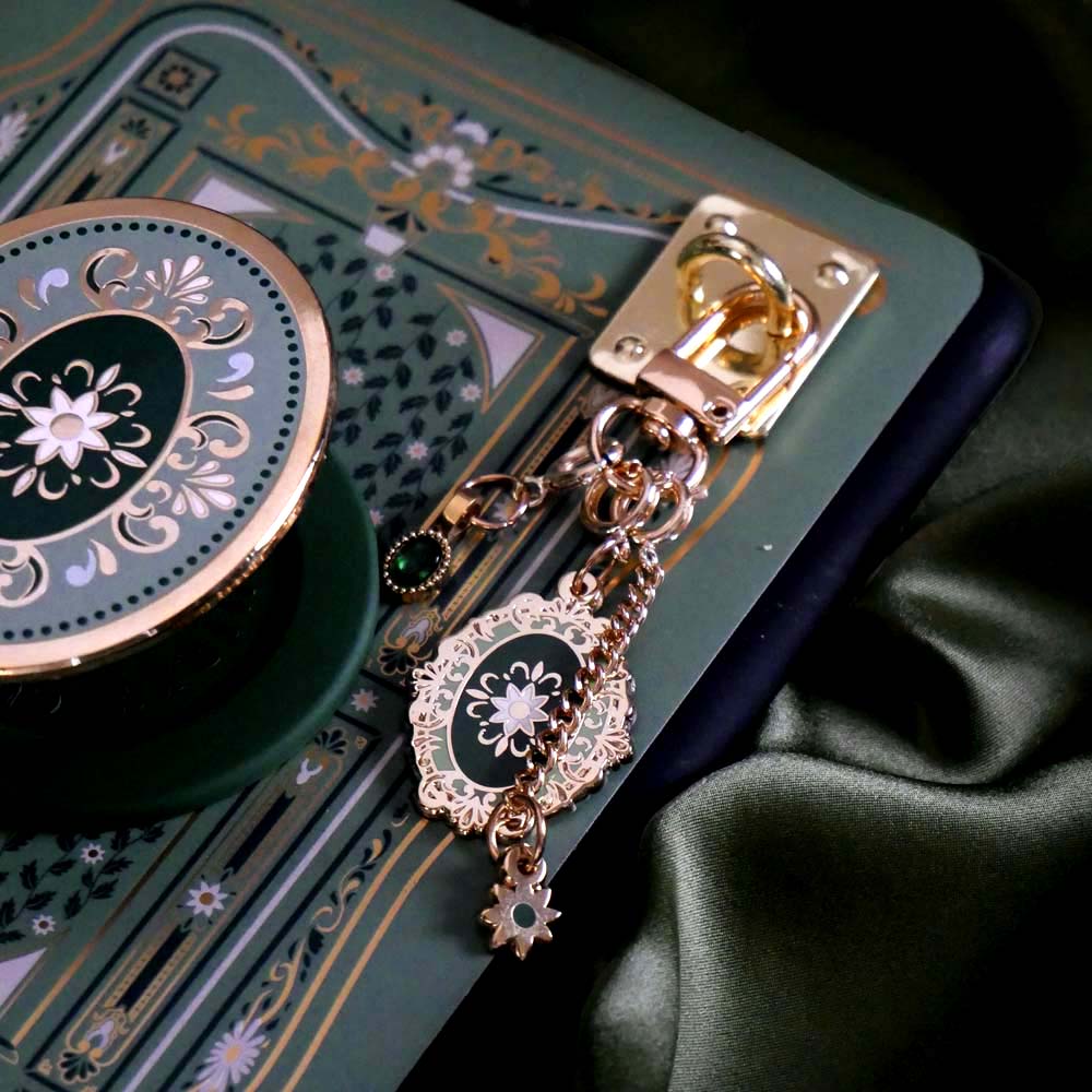The Sage Literati clingie is a gold oval shaped eReader grip with green and gold floral motif design. The kindle grip (similar to an enamel pop socket) is attached to an eReader with a matching green sticker insert and a gold and green Charmlet  all sitting on a sage green background.