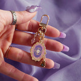 The wisteria literati charmlet by The Quirky Cup Collective is held in a hand with intricate nails in front of a light purple background. The gold charm features three OTHER charms (that can be clipped/ interchanged)- one a small purple gem, one a flower motif on a chain and the main one a pink and purple motif inline with the classical book aesthetic from the Literati collection. 
