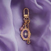 The wisteria literati charmlet by The Quirky Cup Collective sits on a light purple fabric background. The gold charm features three OTHER charms (that can be clipped/ interchanged)- one a small purple gem, one a flower motif on a chain and the main one a pink and purple motif inline with the classical book aesthetic from the Literati collection. 