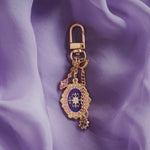 The wisteria literati charmlet by The Quirky Cup Collective sits on a light purple fabric background. The gold charm features three OTHER charms (that can be clipped/ interchanged)- one a small purple gem, one a flower motif on a chain and the main one a pink and purple motif inline with the classical book aesthetic from the Literati collection. 