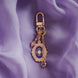 The wisteria literati charmlet by The Quirky Cup Collective sits on a light purple fabric background. The gold charm features three OTHER charms (that can be clipped/ interchanged)- one a small purple gem, one a flower motif on a chain and the main one a pink and purple motif inline with the classical book aesthetic from the Literati collection. 