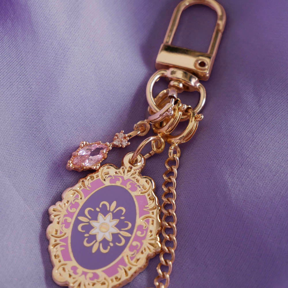 The detail of the wisteria literati charmlet by The Quirky Cup Collective sits on a light purple fabric background. The gold charm features three OTHER charms (that can be clipped/ interchanged)- one a small purple gem, one a flower motif on a chain and the main one a pink and purple motif inline with the classical book aesthetic from the Literati collection. 