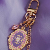 The detail of the wisteria literati charmlet by The Quirky Cup Collective sits on a light purple fabric background. The gold charm features three OTHER charms (that can be clipped/ interchanged)- one a small purple gem, one a flower motif on a chain and the main one a pink and purple motif inline with the classical book aesthetic from the Literati collection. 