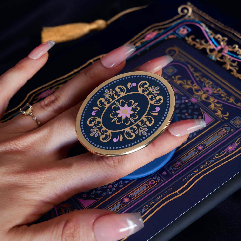 The oval royal blue literati clingie is attached to a kindle with matching eReader sticker insert. The clingie (imagine an enamel pop socket!) features classical book motifs mixed with floral detailing in shades of pastel pink, purple and gold and is held by a hand.