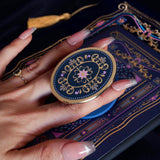 The oval royal blue literati clingie is attached to a kindle with matching eReader sticker insert. The clingie (imagine an enamel pop socket!) features classical book motifs mixed with floral detailing in shades of pastel pink, purple and gold and is held by a hand.