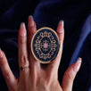 The oval royal blue literati clingie is held in front of a deep blue fabric background. The clingie (imagine an enamel pop socket!) features classical book motifs mixed with floral detailing in shades of pastel pink, purple and gold.