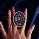 The oval royal blue literati clingie is held in front of a deep blue fabric background. The clingie (imagine an enamel pop socket!) features classical book motifs mixed with floral detailing in shades of pastel pink, purple and gold.