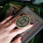 Hand holding a decorative kindle grip with intricate green and purple designs with gold accents on a dark fabric background