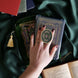 The green Literati clingie by The Quirky Cup Collective is held in front of a matching kindle sticker insert and e-reader sleeve on a green fabric background. The oval shaped gold kindle grip features an intricate classic-style design with pink, green and gold detailing.