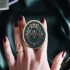 Hand holding a decorative kindle grip with intricate green and purple designs with gold accents on a dark fabric background