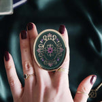 Hand holding a decorative kindle grip with intricate green and purple designs with gold accents on a dark fabric background