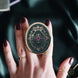 Hand holding a decorative kindle grip with intricate green and purple designs with gold accents on a dark fabric background