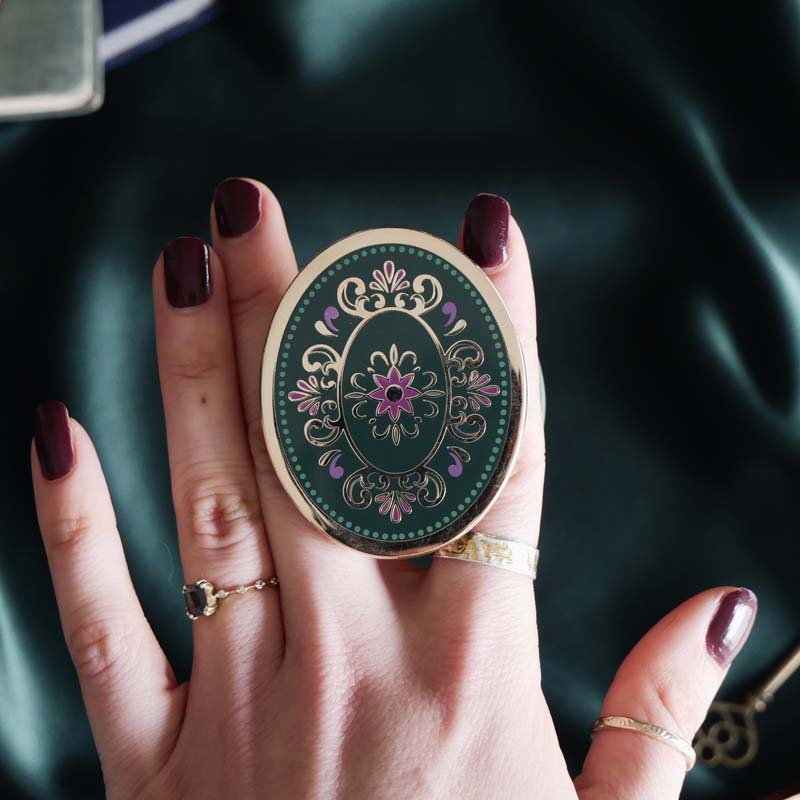Hand holding a decorative kindle grip with intricate green and purple designs with gold accents on a dark fabric background