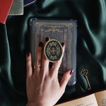 Hand holding a decorative kindle grip with intricate green and purple designs with gold accents on a dark fabric background