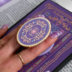 The back of a kindle shows the wisteria light purple literati eReader sticker insert alongside matching oval clingie. Both are light purple and feature pastel pink, purple and blue detailing alongside the gold enamel and foil. The products feature classical Victorian book inspired motifs and the kindle is held in front of a classical book.