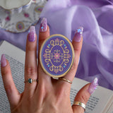 The oval wisteria literati clingie is held in front of a classical book by a hand with intricate purple nails. The clingie (imagine an enamel pop socket!) features classical book motifs mixed with floral detailing in shades of pastel pink, purple and blue mixed with gold. 