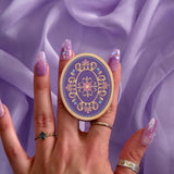 The oval wisteria literati clingie is held by a hand with intricate purple nails in front of a soft purple fabric background. The clingie (imagine an enamel pop socket!) features classical book motifs mixed with floral detailing in shades of pastel pink, purple and blue mixed with gold. 
