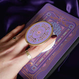 Hand holding a round, decorative clingie MagSafe device grip with intricate designs against a purple fabric background.