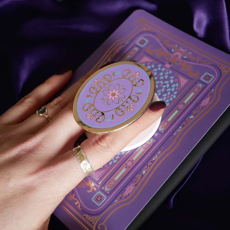 Hand holding a round, decorative clingie MagSafe device grip with intricate designs against a purple fabric background.