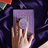 Hand holding a round, decorative clingie MagSafe device grip with intricate designs against a purple fabric background.