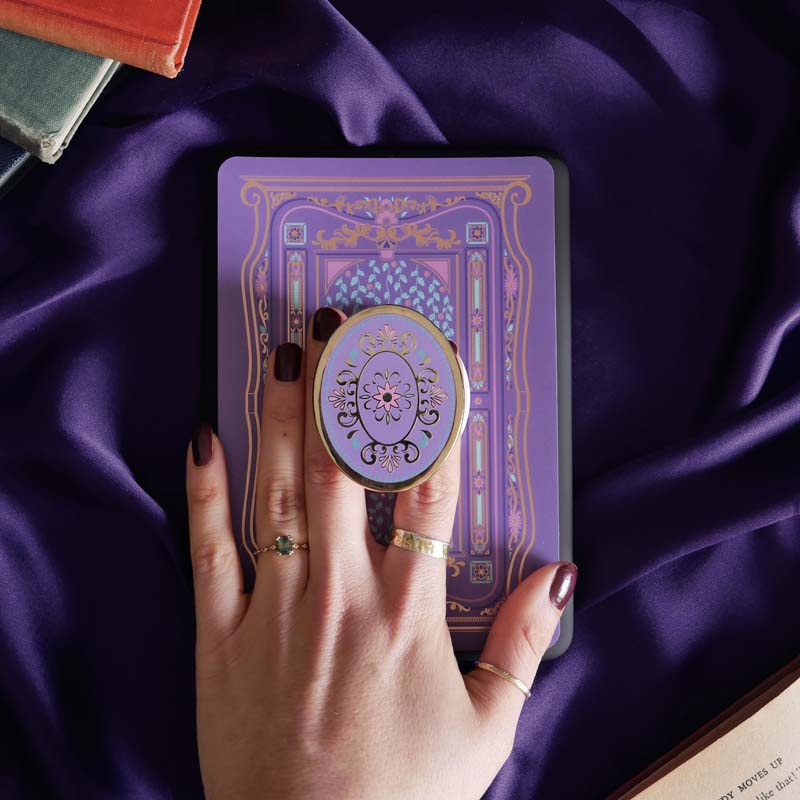Hand holding a round, decorative clingie MagSafe device grip with intricate designs against a purple fabric background.