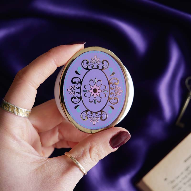 Hand holding a round, decorative clingie MagSafe device grip with intricate designs against a purple fabric background.
