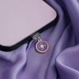 An detail shot of the wisteria literati dust plug in an iphone. The dust plug is light purple with a gold and white flower design in a circle shape. The phone sits on a light purple fabric background.