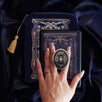 An image of the navy blue literati eReader decor bundle, a hand holding the clingie. The set features an eReader sleeve, eReader sticker insert and clingie all in the same deep royal blue colour with pink and light blue detailing alongside gold foil and embroidery. The intricate victorian inspired design features floral motifs, The eReader case features a gold tassel.