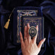 An image of the navy blue literati eReader decor bundle, a hand holding the clingie. The set features an eReader sleeve, eReader sticker insert and clingie all in the same deep royal blue colour with pink and light blue detailing alongside gold foil and embroidery. The intricate victorian inspired design features floral motifs, The eReader case features a gold tassel.