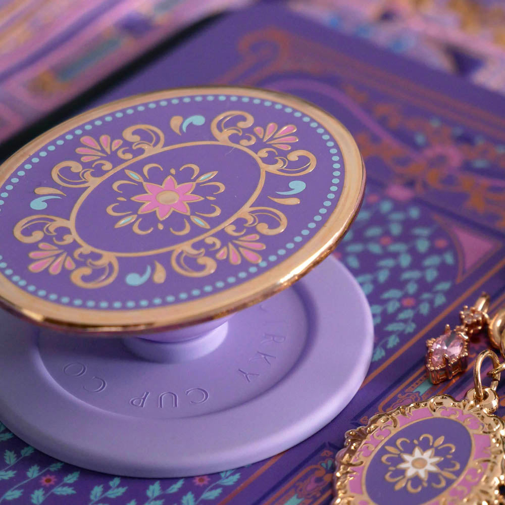 A detail of the wisteria light purple literati eReader sticker insert alongside matching oval clingie and charmlet. Both are light purple and feature pastel pink, purple and blue detailing alongside the gold enamel and foil. The products feature classical Victorian book inspired motifs. The clingie features the brand logo on the back.