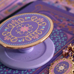 A detail of the wisteria light purple literati eReader sticker insert alongside matching oval clingie and charmlet. Both are light purple and feature pastel pink, purple and blue detailing alongside the gold enamel and foil. The products feature classical Victorian book inspired motifs. The clingie features the brand logo on the back.