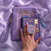An image of the wisteria literati eReader decor bundle, a hand holding the clingie. The set features an eReader sleeve, eReader sticker insert, charmlet and dust plug all in the same light purple colour with pastel pink, purple and blue detailing alongside gold foil and embroidery. The intricate victorian inspired design features floral motifs, The eReader case features a wrist strap and blue tassel.