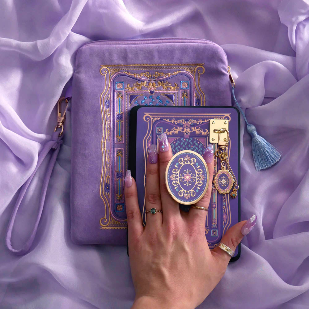An image of the wisteria literati eReader decor bundle, a hand holding the clingie. The set features an eReader sleeve, eReader sticker insert, charmlet and dust plug all in the same light purple colour with pastel pink, purple and blue detailing alongside gold foil and embroidery. The intricate victorian inspired design features floral motifs, The eReader case features a wrist strap and blue tassel.