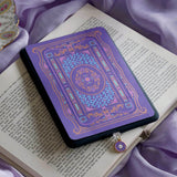 An detail shot of the wisteria literati eReader insert sticker on a kindle alongside the matching dust plug. The insert sticker is light purple and features intricate victorian motifs of flowers and books in shades of pink, purple, blue and foiled gold. The dust plug is light purple with a gold and white flower design in a circle shape. The kindle sits on an open classical book on a  light purple fabric background. 