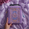 The lilac Literati eReader sleeve in Wisteria sits on a light purple fabric background. The velvet kindle case features a classical design inspired by victorian classical books with detailed embroidery in shades of blue, purple and gold. The case also features a wrist strap and a red light blue tassel.