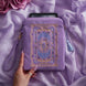 The lilac Literati eReader sleeve in Wisteria sits on a light purple fabric background. The velvet kindle case features a classical design inspired by victorian classical books with detailed embroidery in shades of blue, purple and gold. The case also features a wrist strap and a red light blue tassel.