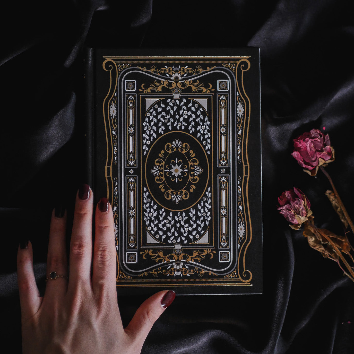 A hand holding the Black Literati Journal by The Quirky Cup Collective, showcasing its intricate gold and white floral and geometric cover design on a black background. The journal is positioned on top of a dark, silky fabric, with dried pink roses placed next to it, creating a moody, vintage-inspired scene. The journal's ribbon bookmark is partially visible, adding a touch of elegance to the overall aesthetic.