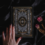 A hand holding the Black Literati Journal by The Quirky Cup Collective, showcasing its intricate gold and white floral and geometric cover design on a black background. The journal is positioned on top of a dark, silky fabric, with dried pink roses placed next to it, creating a moody, vintage-inspired scene. The journal's ribbon bookmark is partially visible, adding a touch of elegance to the overall aesthetic.