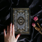 A hand holding the Black Literati Journal by The Quirky Cup Collective, showcasing its intricate gold and white floral and geometric cover design on a black background. The journal is positioned on top of a dark, silky fabric, with dried pink roses placed next to it, creating a moody, vintage-inspired scene. The journal's ribbon bookmark is partially visible, adding a touch of elegance to the overall aesthetic.