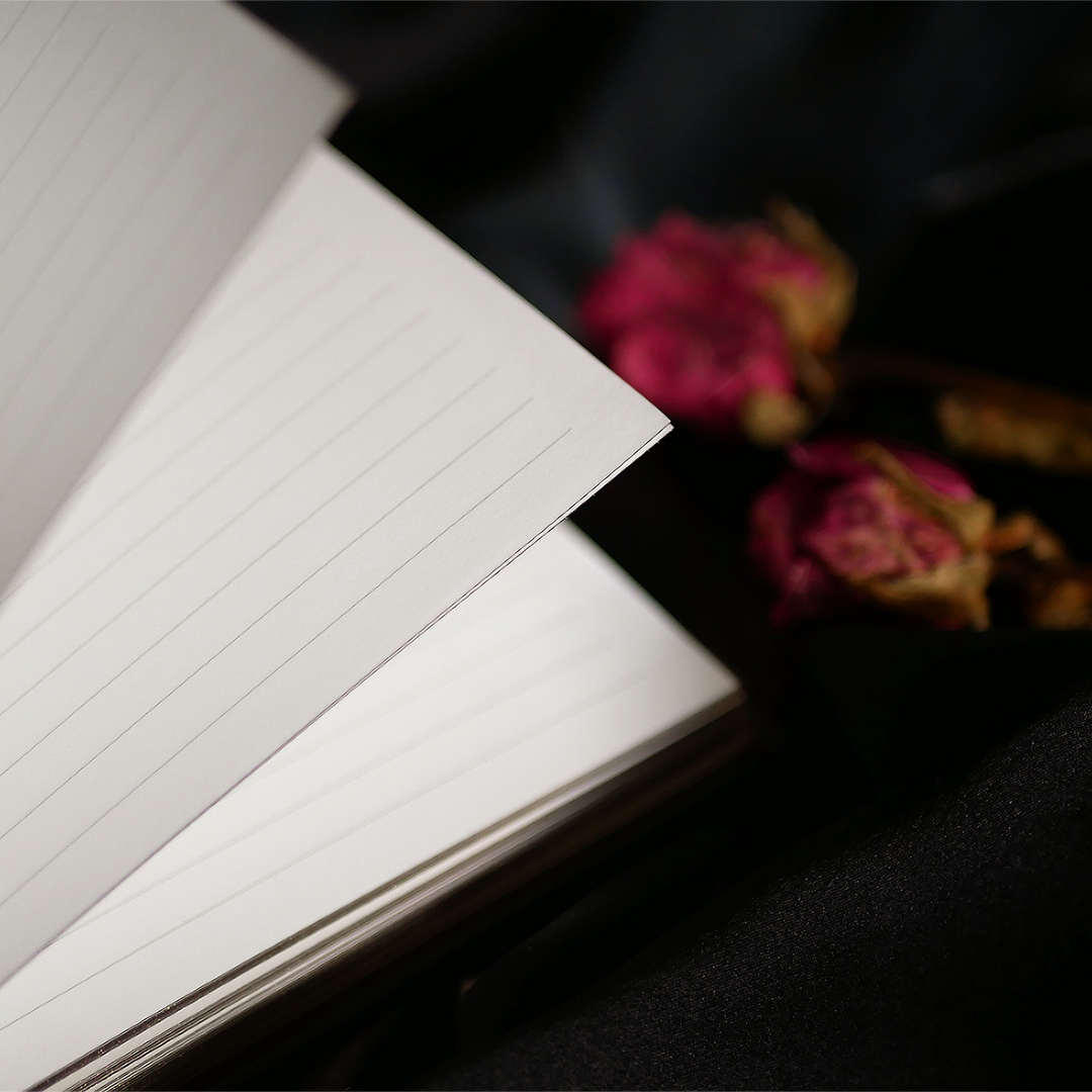 A close-up of a Black Literati Journal by The Quirky Cup Collective, featuring a patterned cover with an ornate design. A hand with dark red nail polish is slightly blurred in the foreground, suggesting movement or anticipation. The journal rests on a dark fabric background, with dried pink roses placed to the side, adding a touch of vintage elegance to the composition.